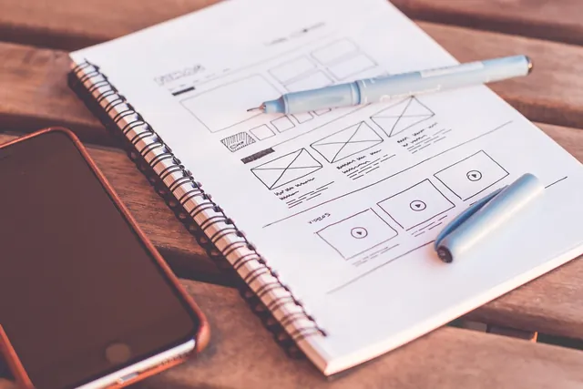 Wireframe de um site esboçado num caderno, ao lado de um telemóvel